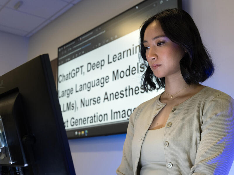 A photo of a woman from the chest up. She is standing behind a computer monitor and in front of a display screen. The screen has black text that reads "ChatGPT, Deep Learning Large Language Models (LMs), Nurse Anesthetics, Next Generation Image"
