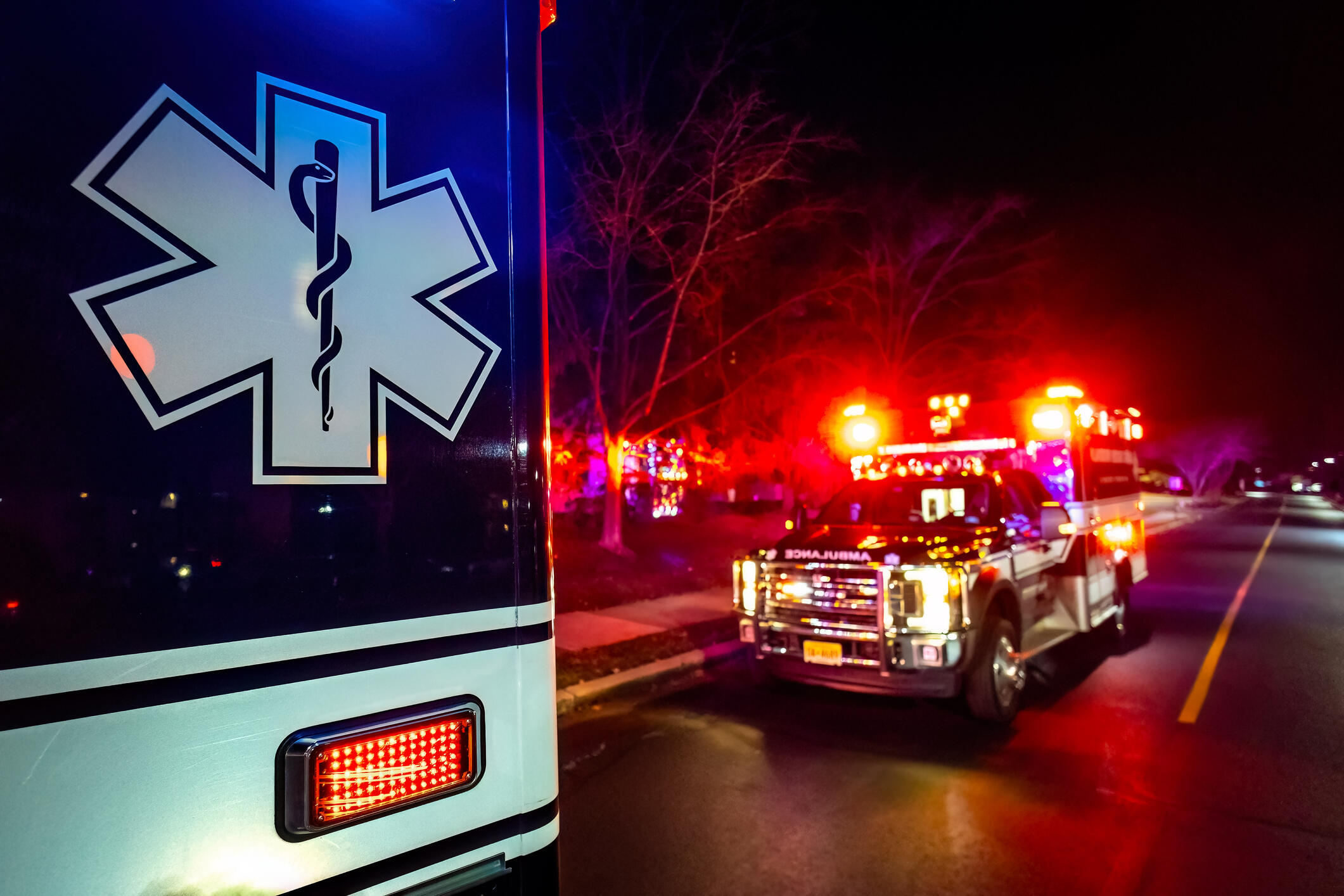 A photo of an ambulance up close and one parked behind it. The ambulance in the back has its emergency lights on. 