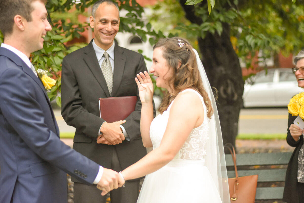 A bride and groom hold hands and laugh while standing in front of an officiant, who is also laughing.