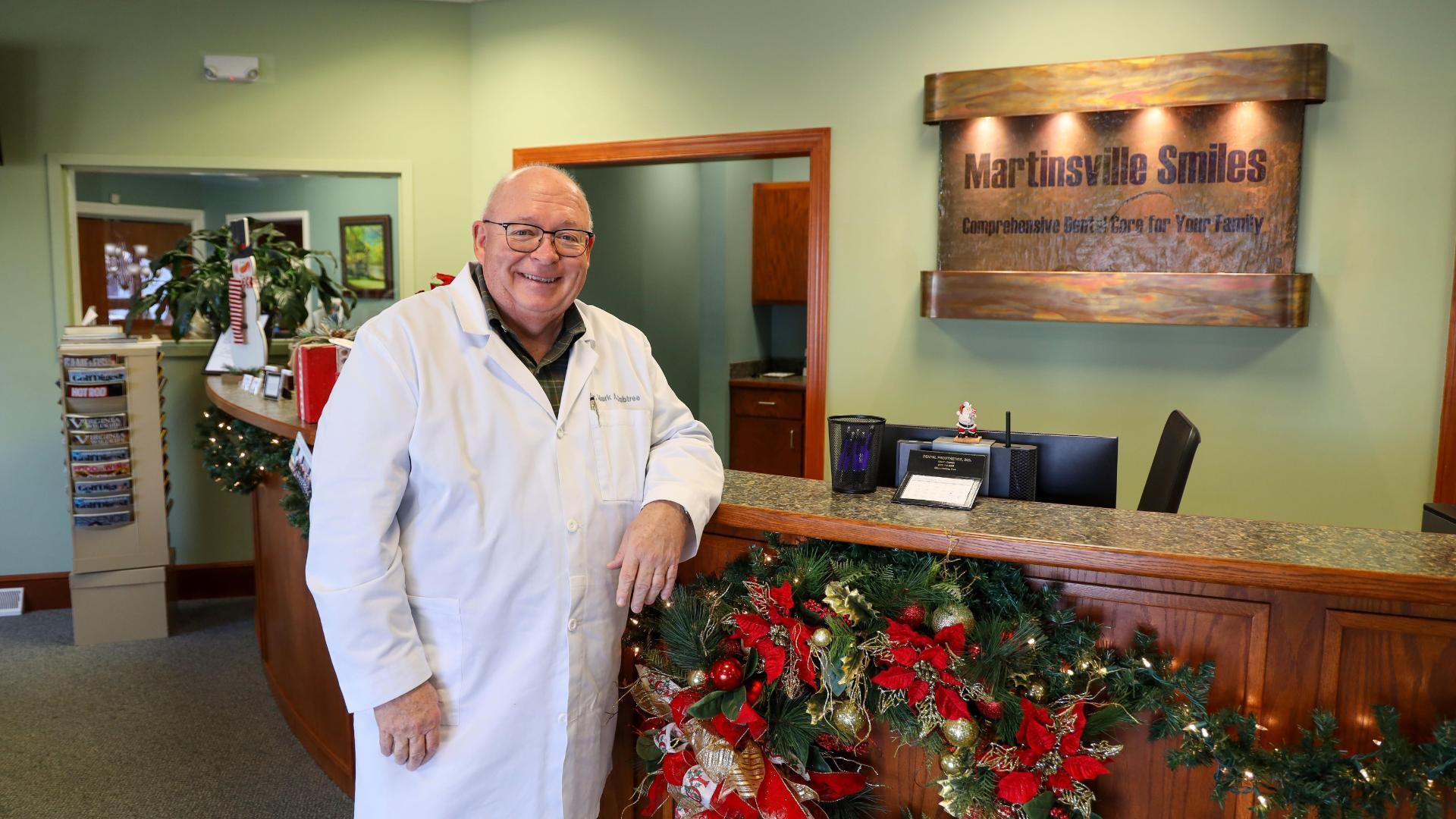 A photo of a man wearing a white medical coat standing next to a check in desk. 