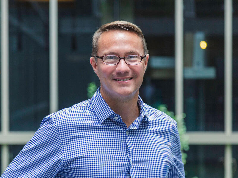 A photo of a man from the chest up wearing a blue and white button down shirt and black glasses. 