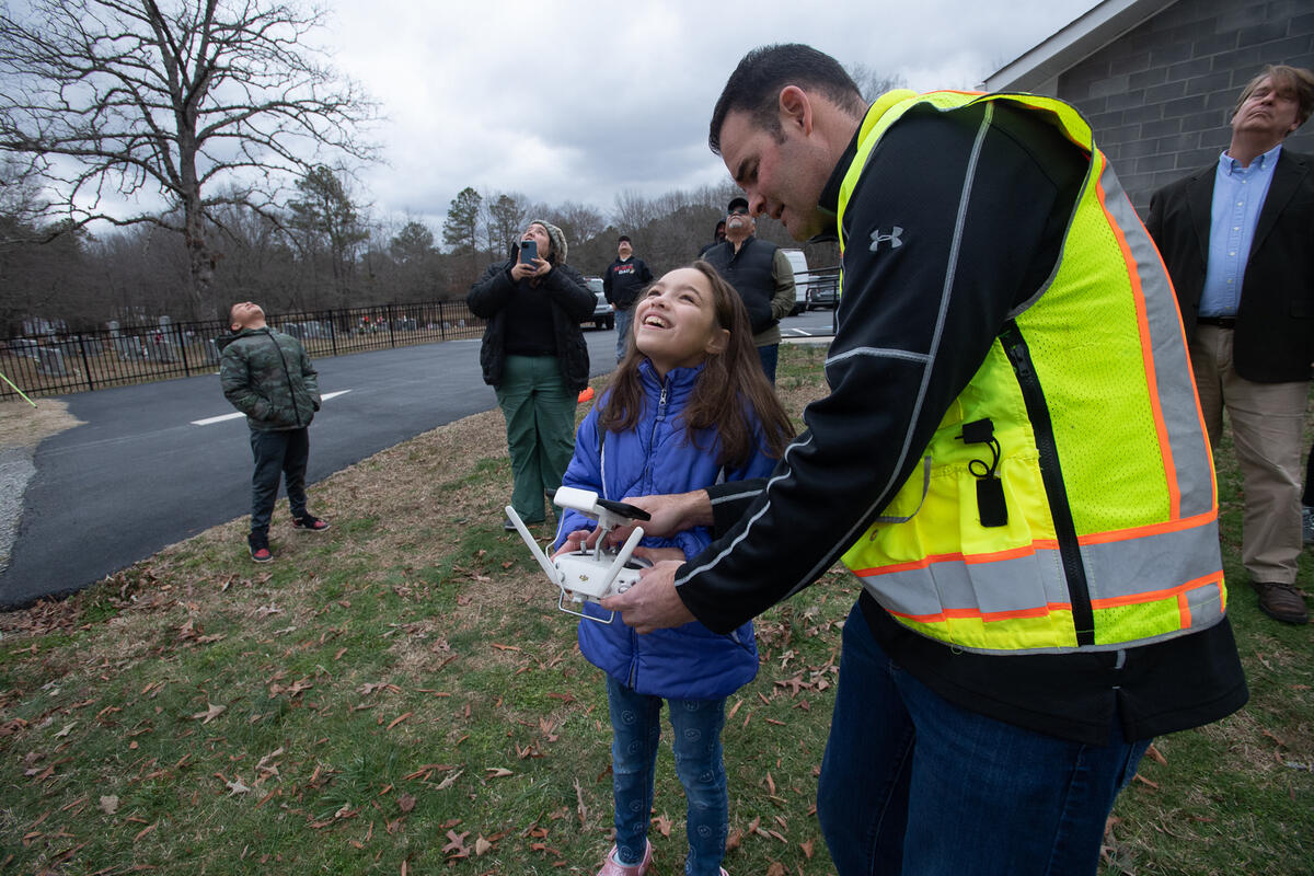 A photo of a man and a young girl holding the remote for a dorone. The man is looking down while the girl looks up at the sky. There are five people around them also looking up at the sky. 