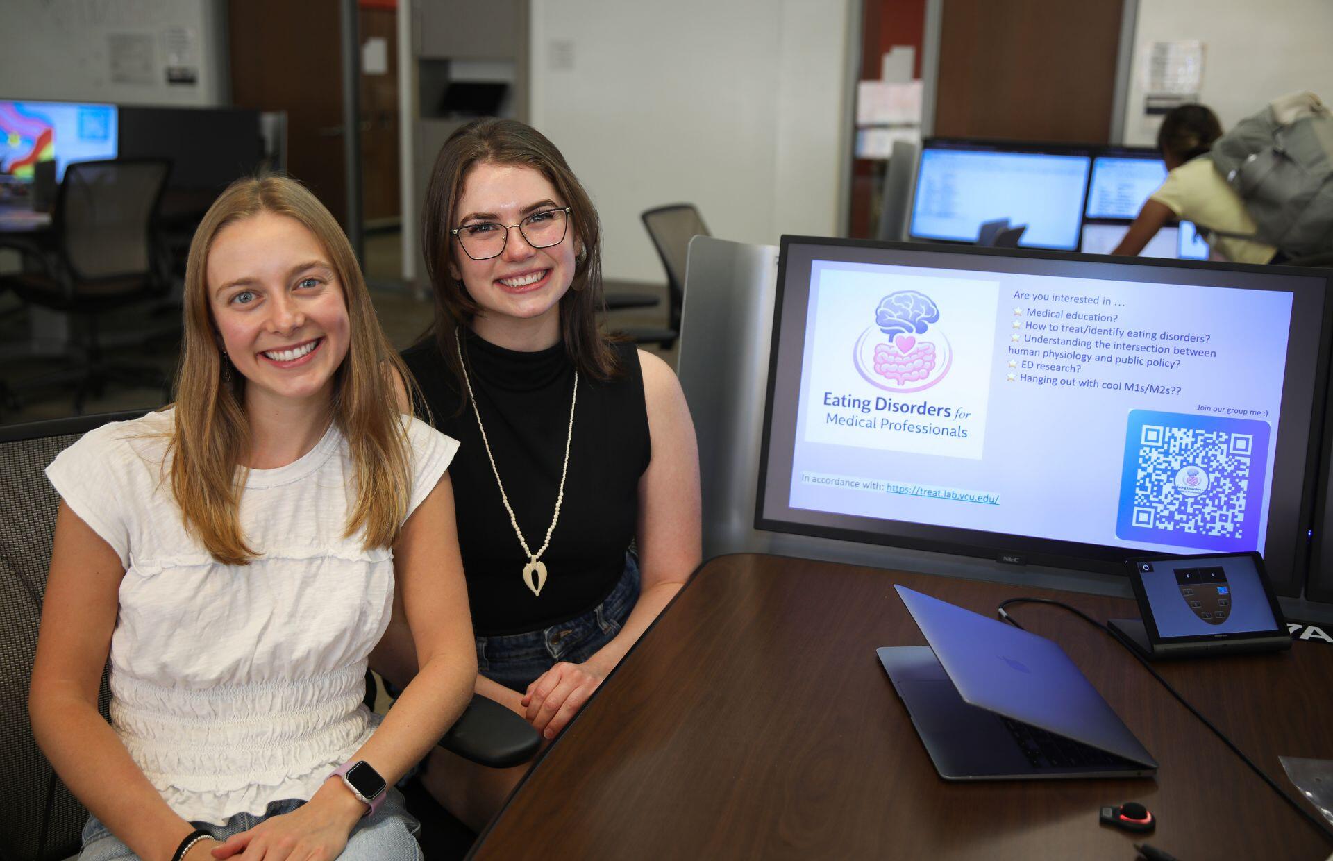 A photo of two women sitting to the left of a table with a laptop and a computer monitor on it.
