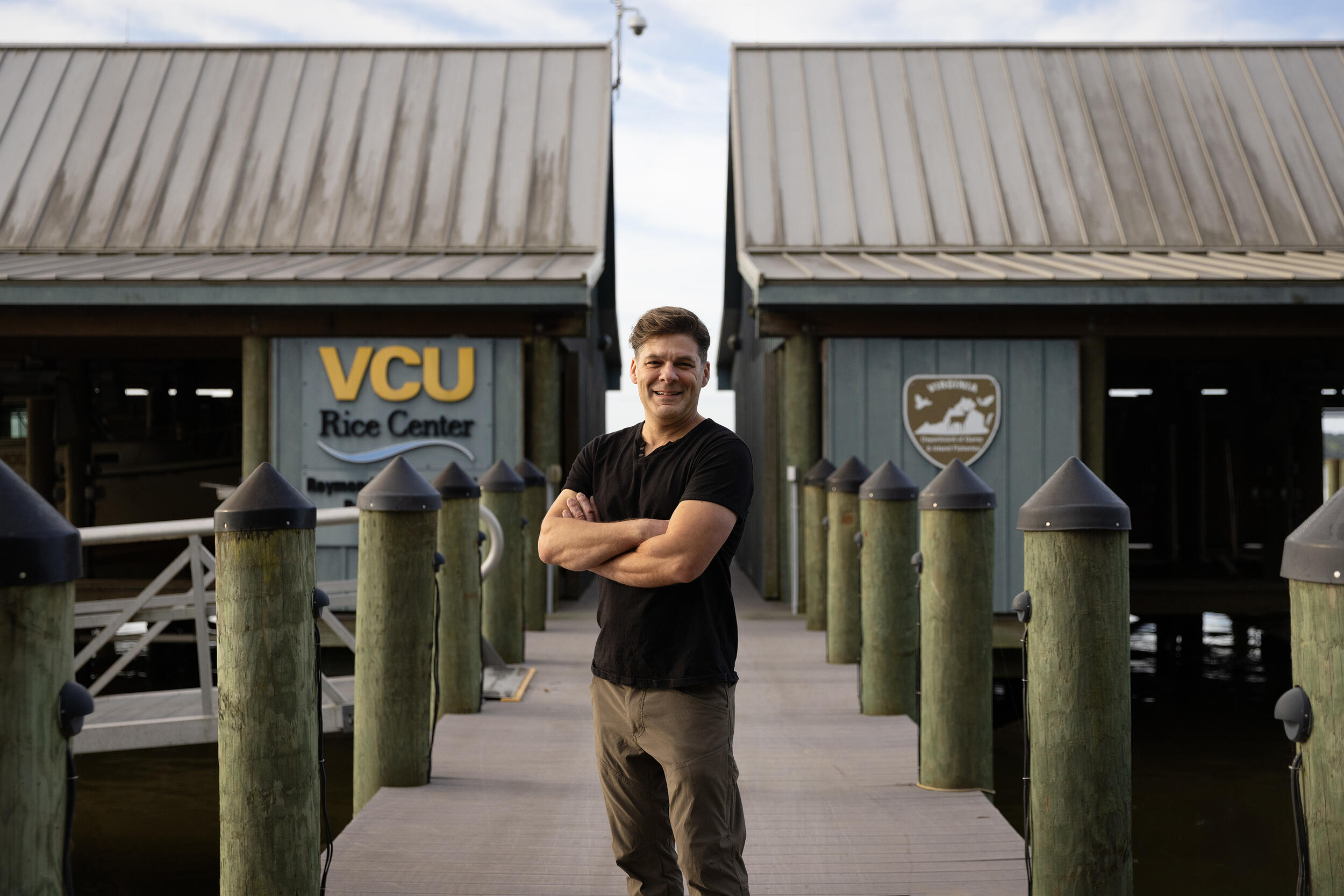 A photo of a man standing on a dock with his arms crossed against his chest. Behind him are two buildings. The building on the left has a sign that reads \"VCU Rice Center.\" 