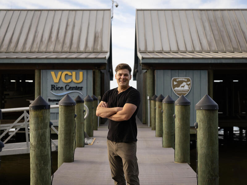 A photo of a man standing on a dock with his arms crossed against his chest. Behind him are two buildings. The building on the left has a sign that reads \"VCU Rice Center.\" 
