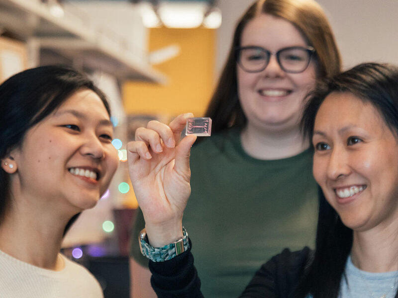 Three women next to eachother. They are all looking at a small clear rectangle with a small object in it. All of the women are smiling and looking at the object. 