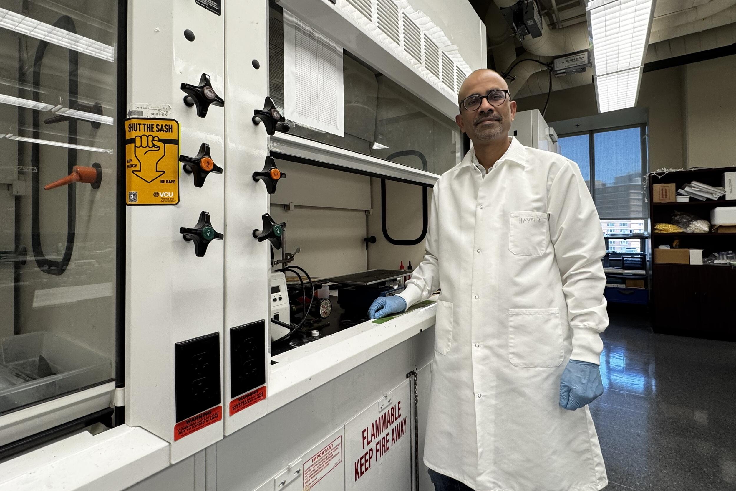 A photo of a man in a white lap coat, blue latex gloves, and safety glasses standing next to a fume hood in a science lab. 