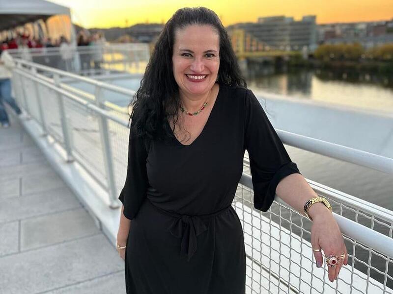 A photo of a woman standing next to a fence, with her right arm resting on a railing. Behind her is a river and buildings.
