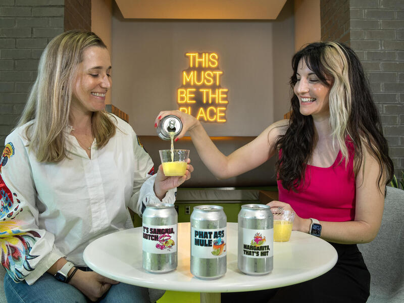 A photo of two women sitting at a small circular table. The woman on the right is pouring a can with organge liquid into the woman on the left's cup. 