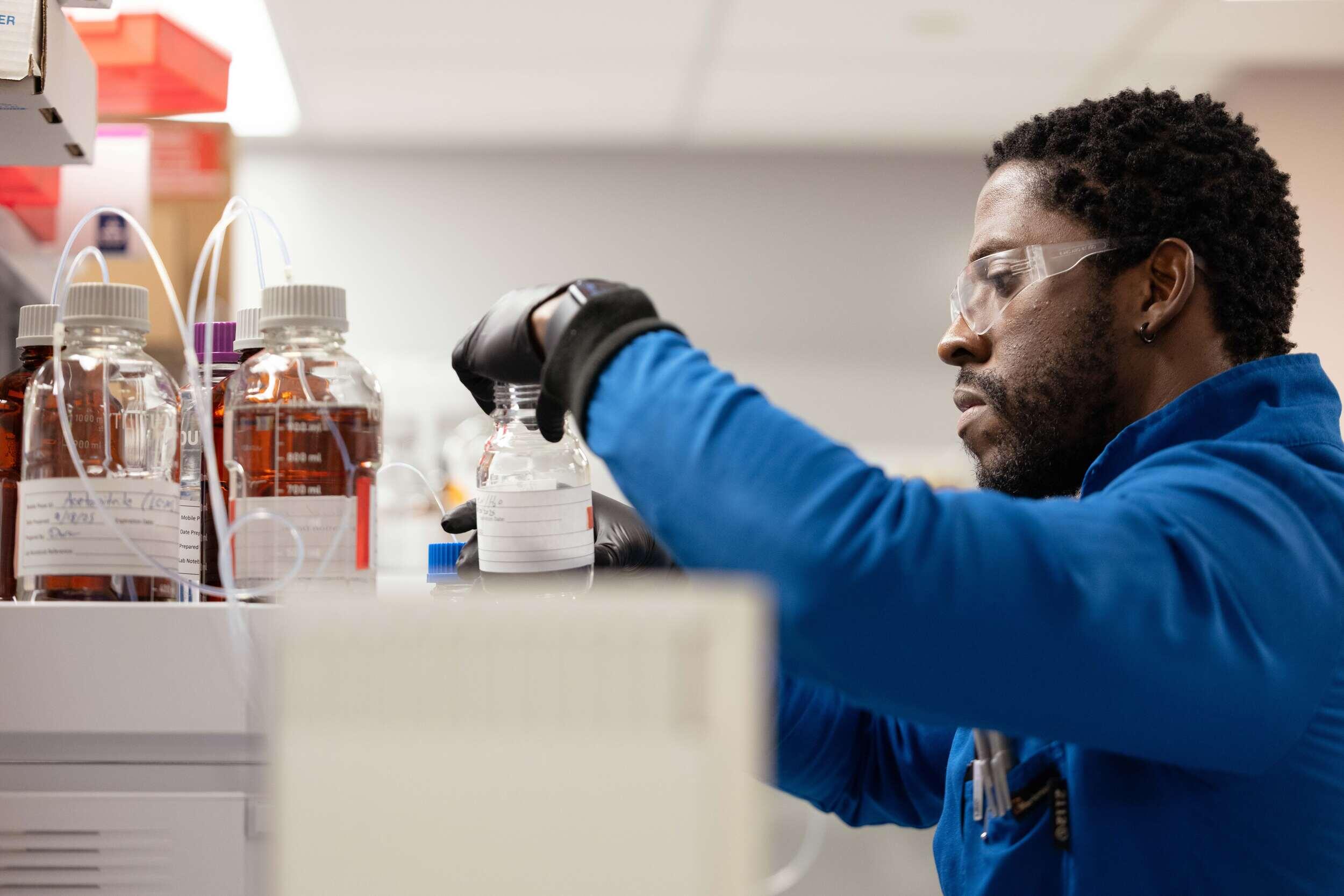 A photo of a man in a lab wearing a blue lab coat, black latex gloves, and safety glasses, holding a glass bottle. 