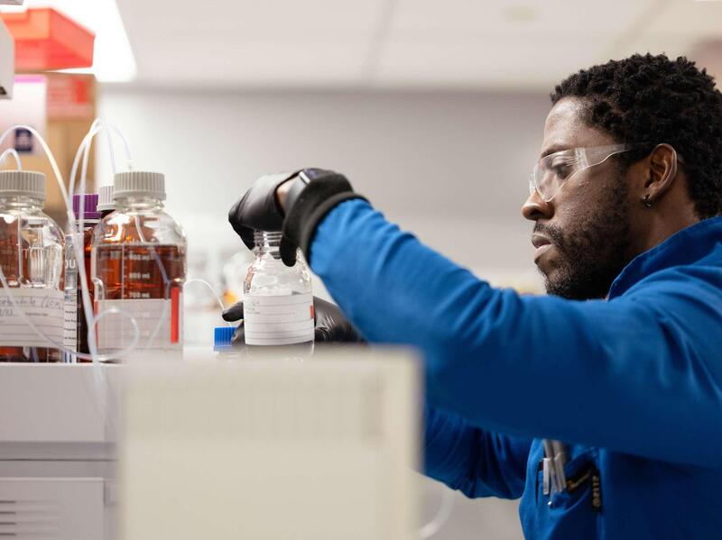 A photo of a man in a lab wearing a blue lab coat, black latex gloves, and safety glasses, holding a glass bottle. 