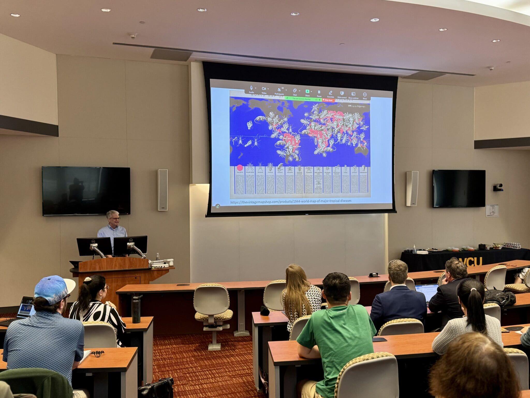 A photo of a college classroom. Students are seated at desks looking up a the front of the room. At the front of the room is a man standing behind a podium with two computer monitors on it. To the right of the man is a screen with an image of a map being projected on it. 