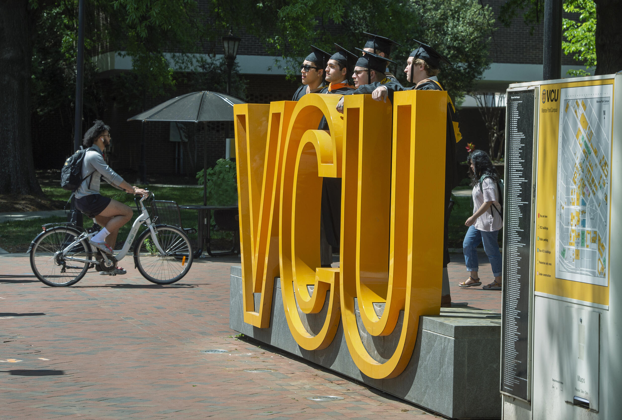 A photo of three people wearing graduation cap and gowns standing behind a sign made of big yellow letters that spell out \"VCU\"