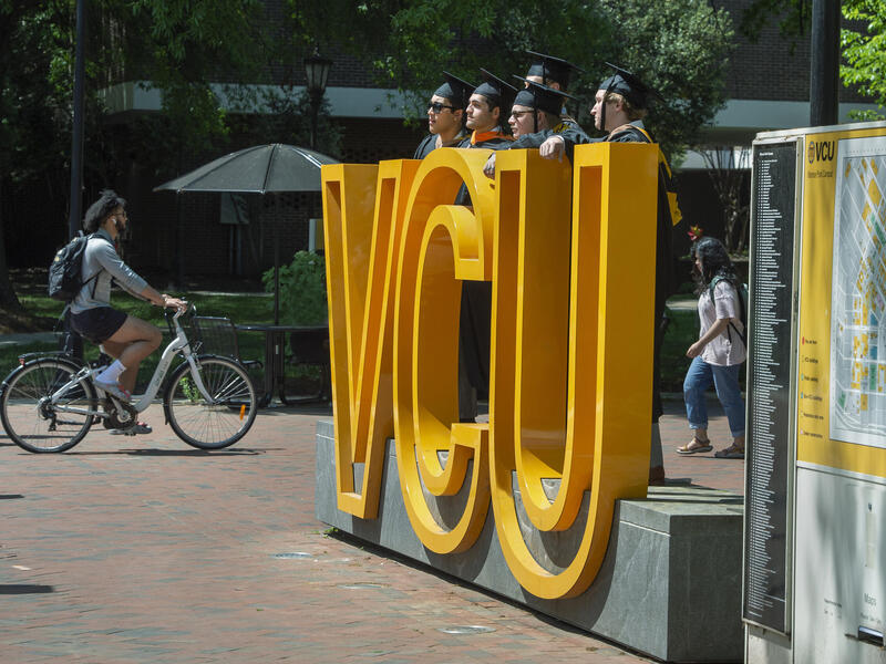 A photo of three people wearing graduation cap and gowns standing behind a sign made of big yellow letters that spell out \"VCU\"