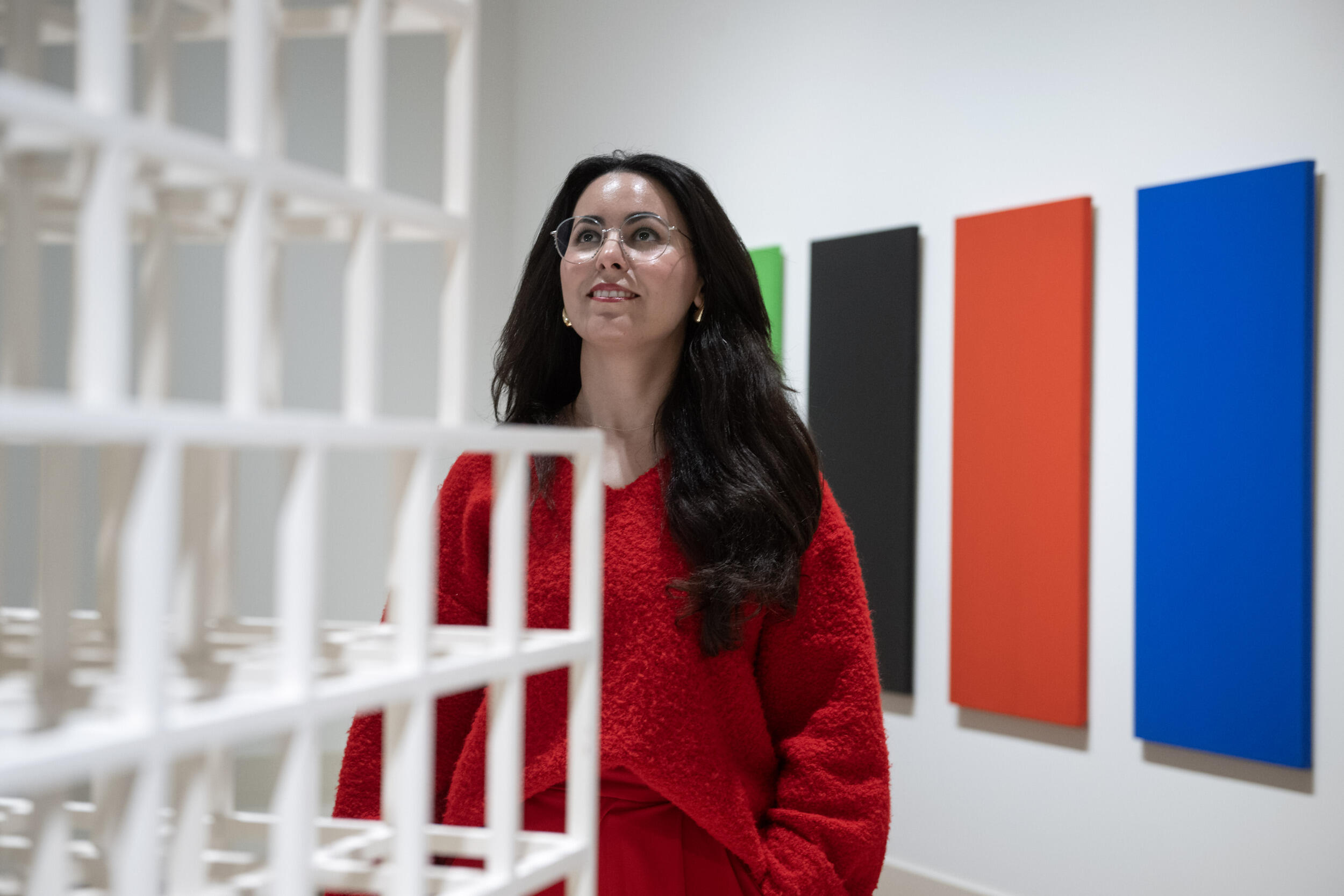 A photo of a woman standing in front of a white sculpture. Behind her are four rectangular green, black, red, and blue canvases. 