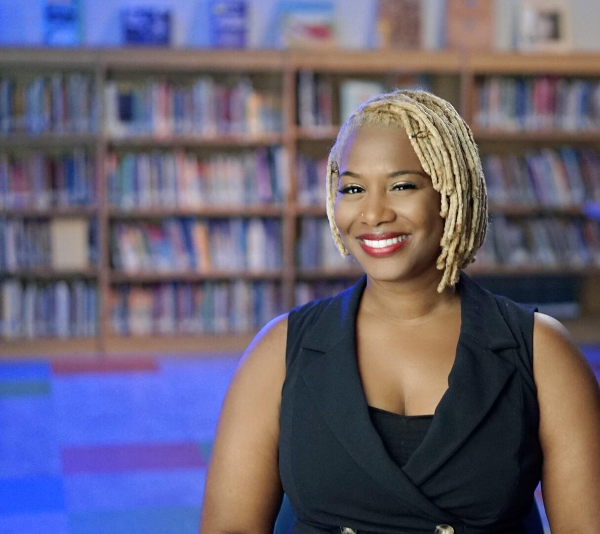 A portait of a woman from the chest up standing in front of a bookshelf. 