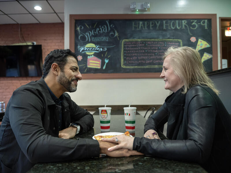 A photo of a man and a woman sitting across from each other at a table. They are looking at each other lovingly and the woman has her hand on one of the man's. 