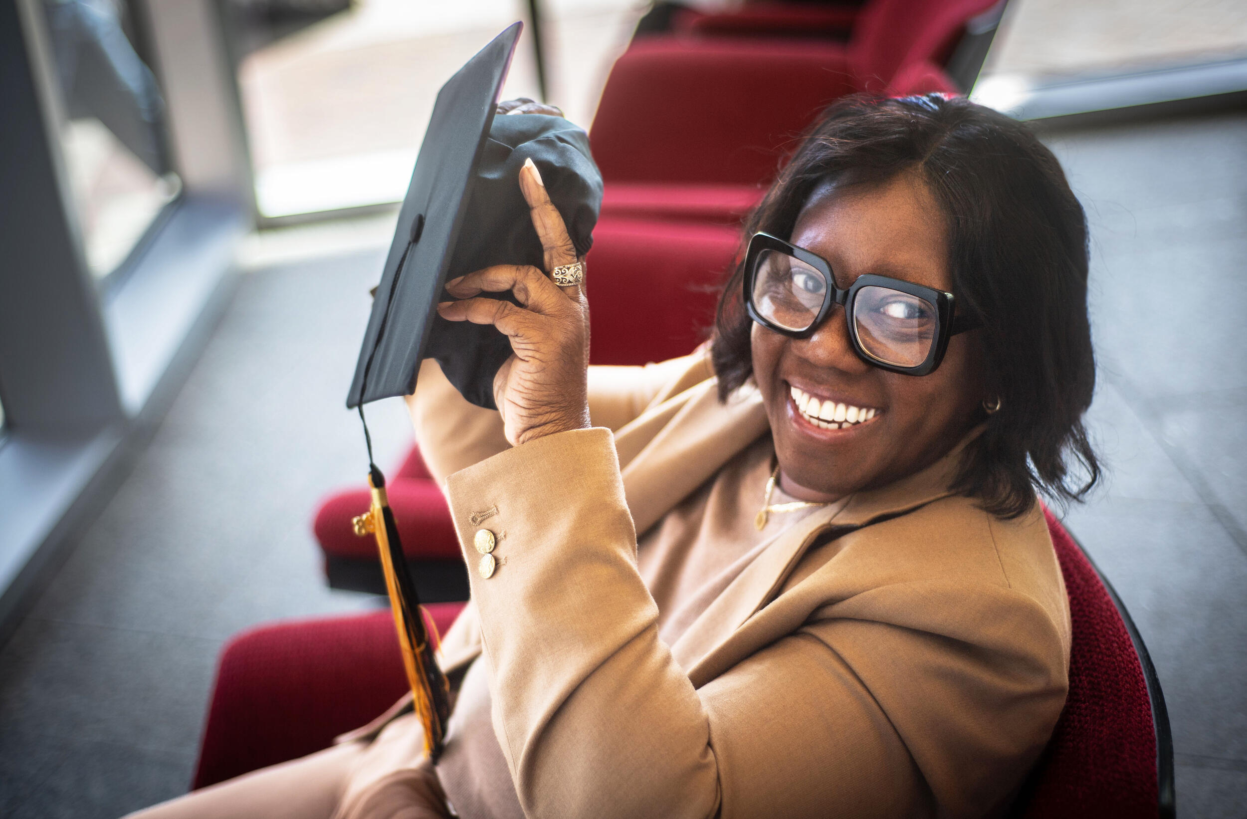A photo of a a woman sitting in a red chair. She is smiling and holding a black graduation cap. 