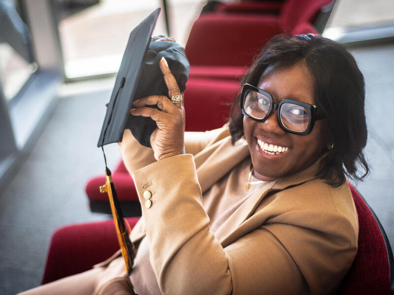 A photo of a a woman sitting in a red chair. She is smiling and holding a black graduation cap. 
