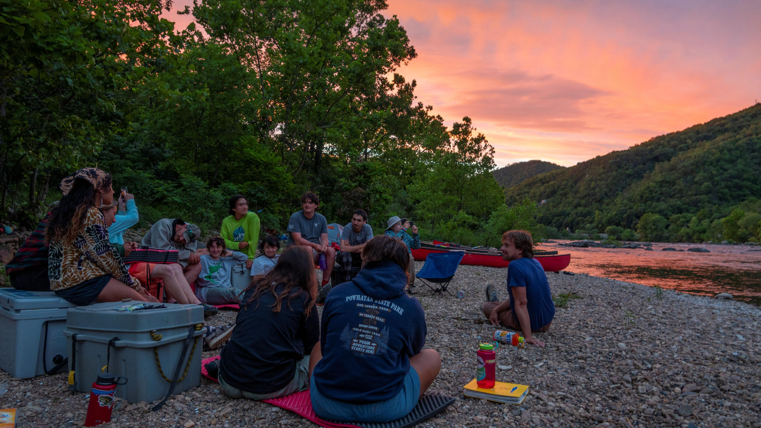 A group of men and women sit on the rocky shore of a river at dusk.