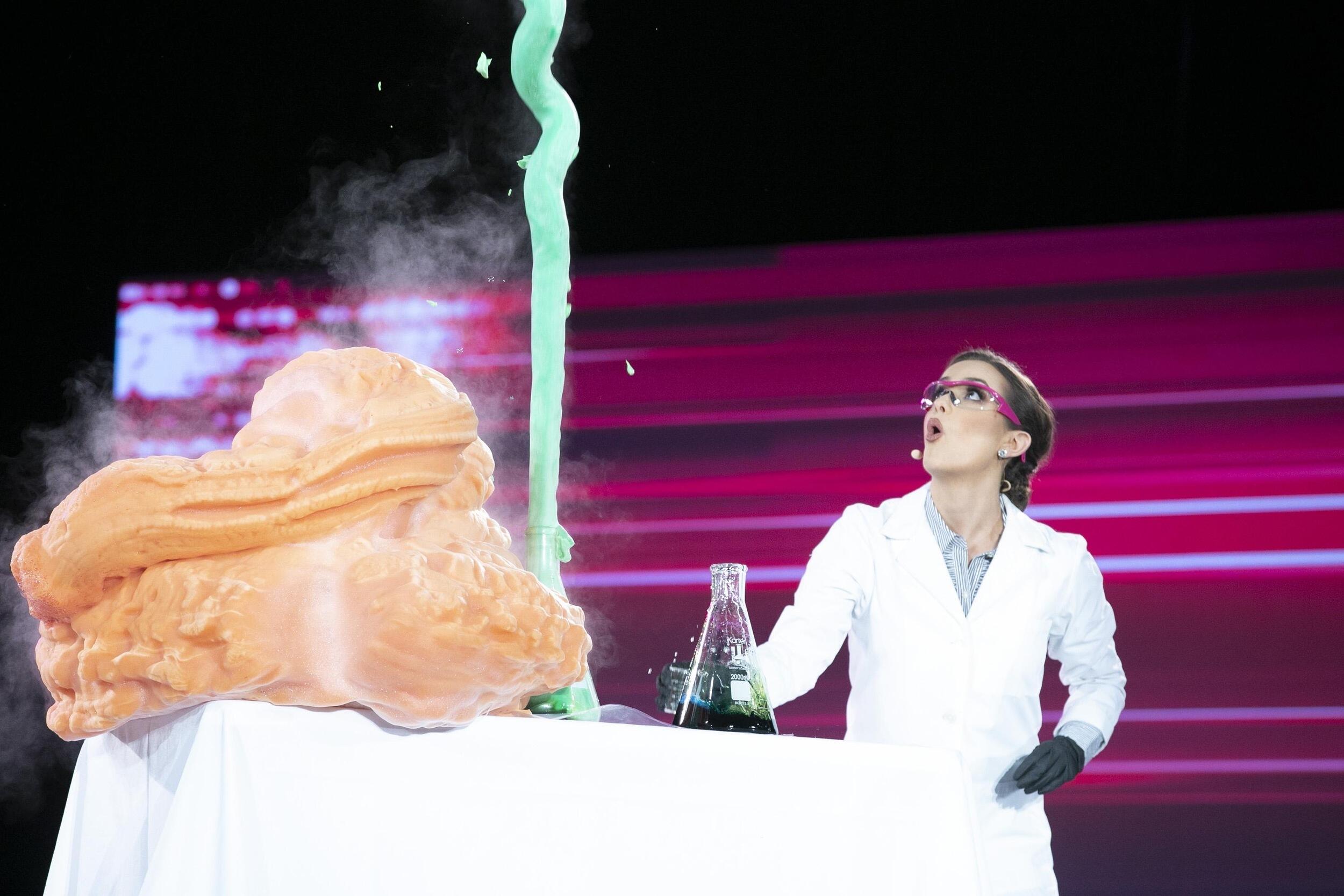 A photo of a woman wearing a labcoat looking up at a green susbstance shooting out of a beaker on a table. 