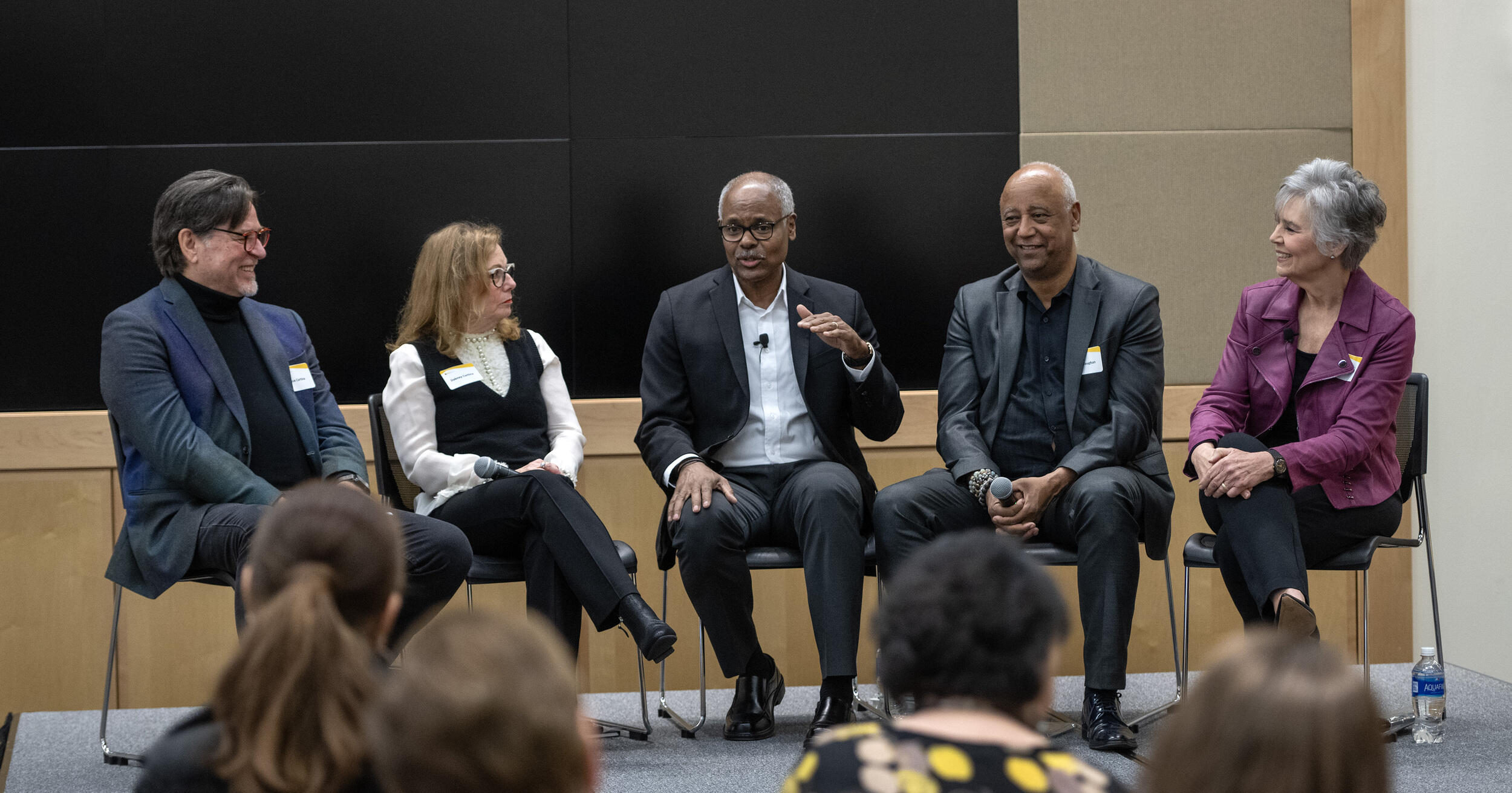 A photo of five people sitting on a stage. The man in the middle is speaking while the four people next to him listen. 