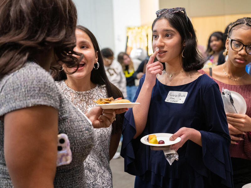A photo of four women standing. Two are holding plates with food and one is looking at their phone. 
