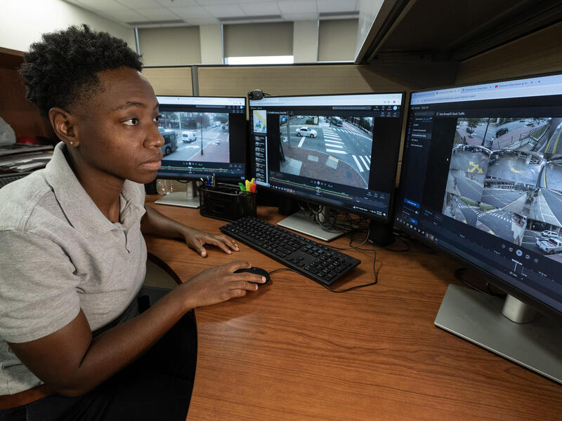 A photo of a person sitting at a desk with three computer monitors on it. The person has their hand on a computer mouse and is looking at the far right screen. Each computer screen has feed from security cameras on it. 