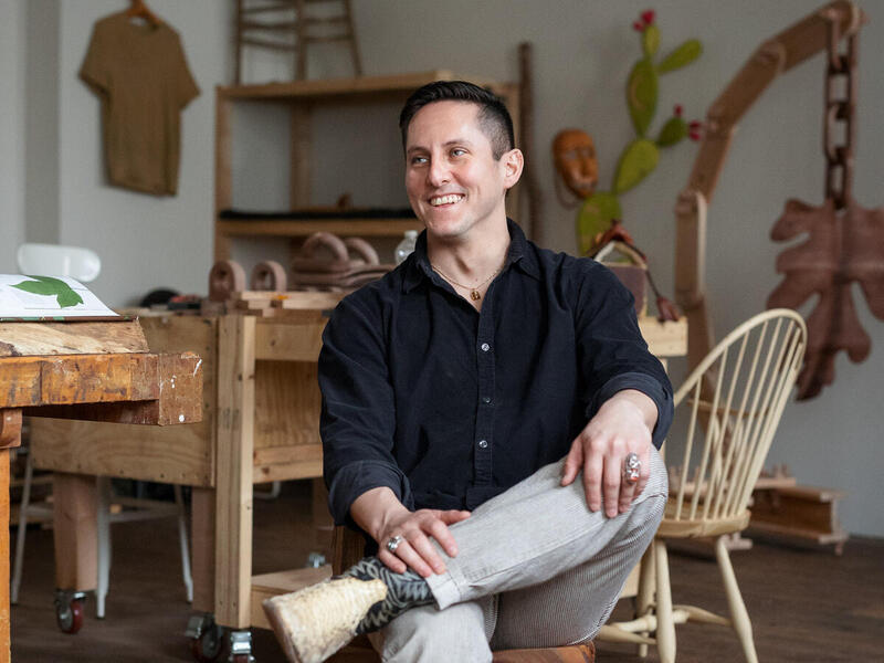 Man sits in a chair with his legs crossed and smiles looking off camera.