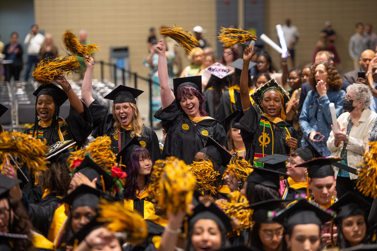 A photo of a crowd of people wearing graduation caps and gowns cheering and waving pompoms in the air. 