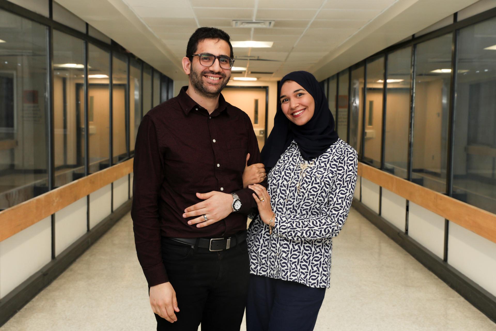 A photo of a mand and a woman standing and smiling. They are holding arms and standing in a hallway. 