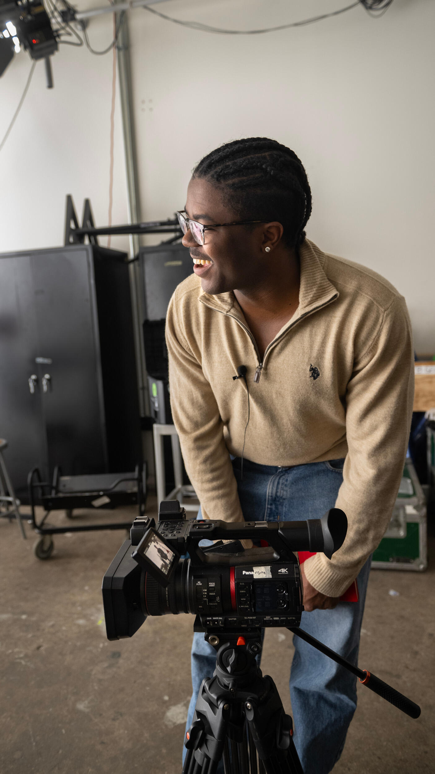 A man smiles and leans over a video camera, looking offscreen to the left.