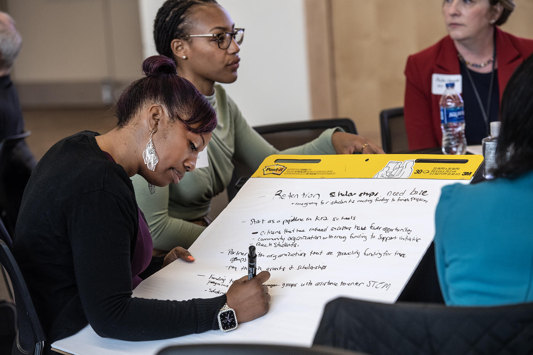 Four people sitting at a table. One of the woman is writing on a giant pad of paper. 