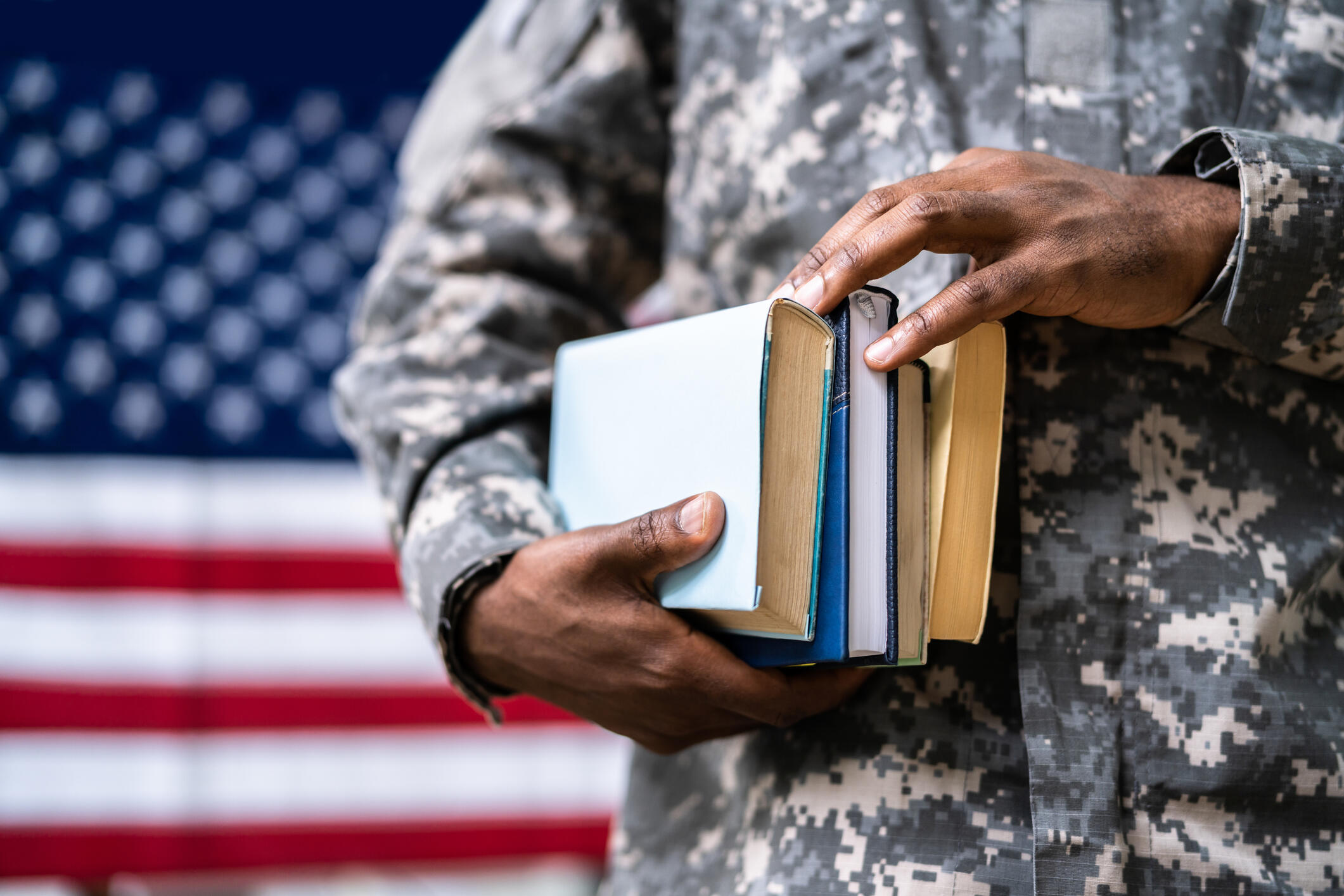 A photo of a person in a military uniform from the shoulders down. They are holding a stack of four books and standing in front of an American flag. 