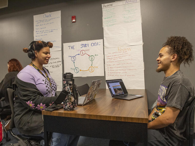 A photo of two people sitting across from each other at a table with laptops sitting on the table 
