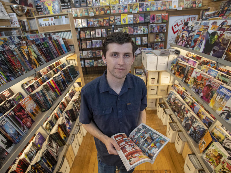 A man standing in the middle of a comic shop holding an open comic book. All around him are shelves lined with comic books and graphic novels. 