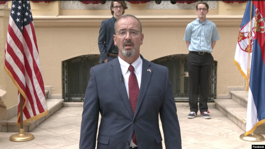 A photo of a man wearing a suit from the chest up. There are two other men standing behind him. 