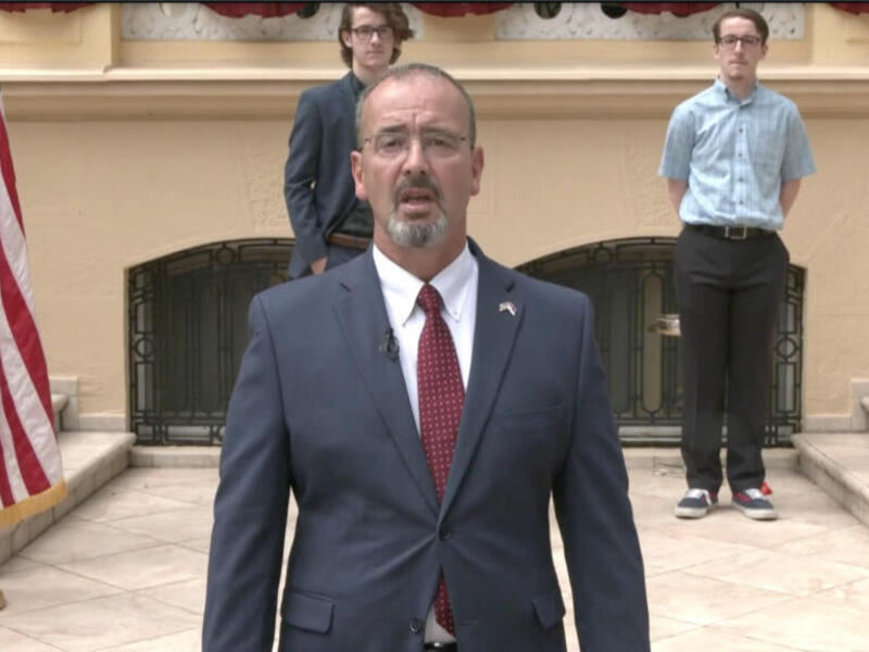 A photo of a man wearing a suit from the chest up. There are two other men standing behind him. 