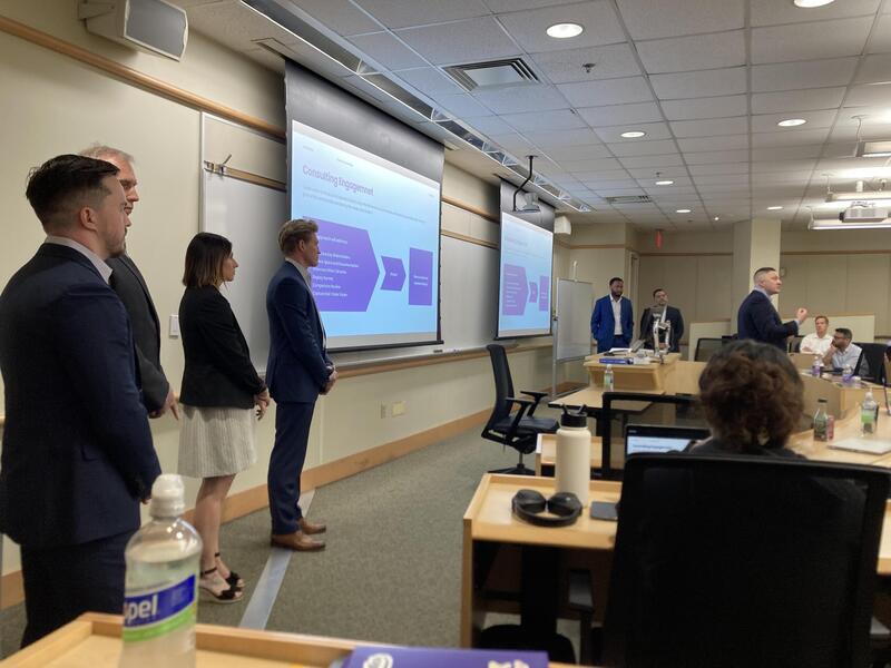 A photo of six people standing at the front of a classroom with a seventh person standing slighting in front of them talking. To the sides of the room, three people are sitting and looking at the person in the front speaking. 