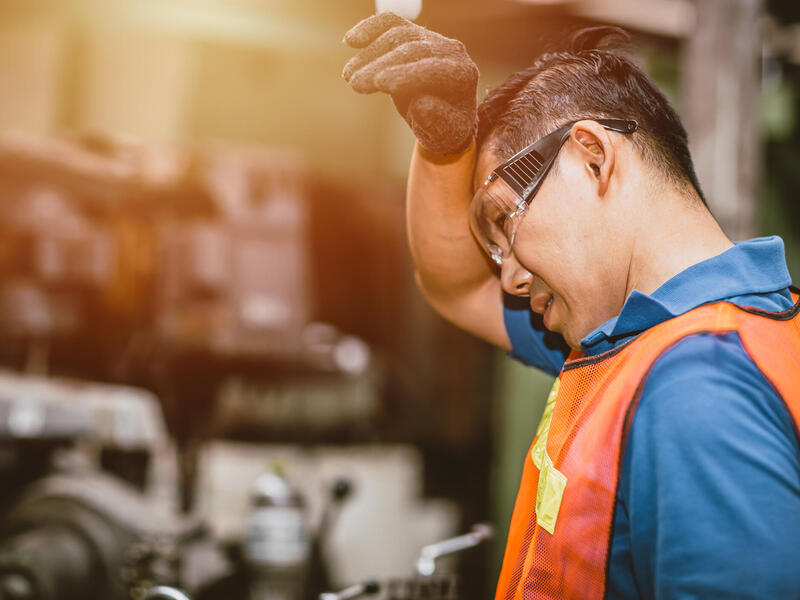 A man wearing a blue collar shirt and orange vest wiping his forhead with his arm. 