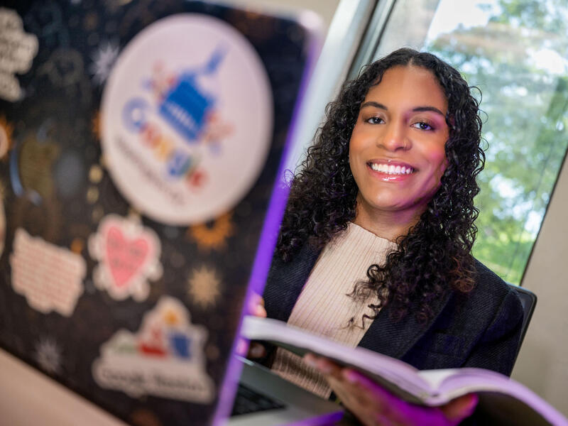 A woman holds an open book in front of an open laptop.