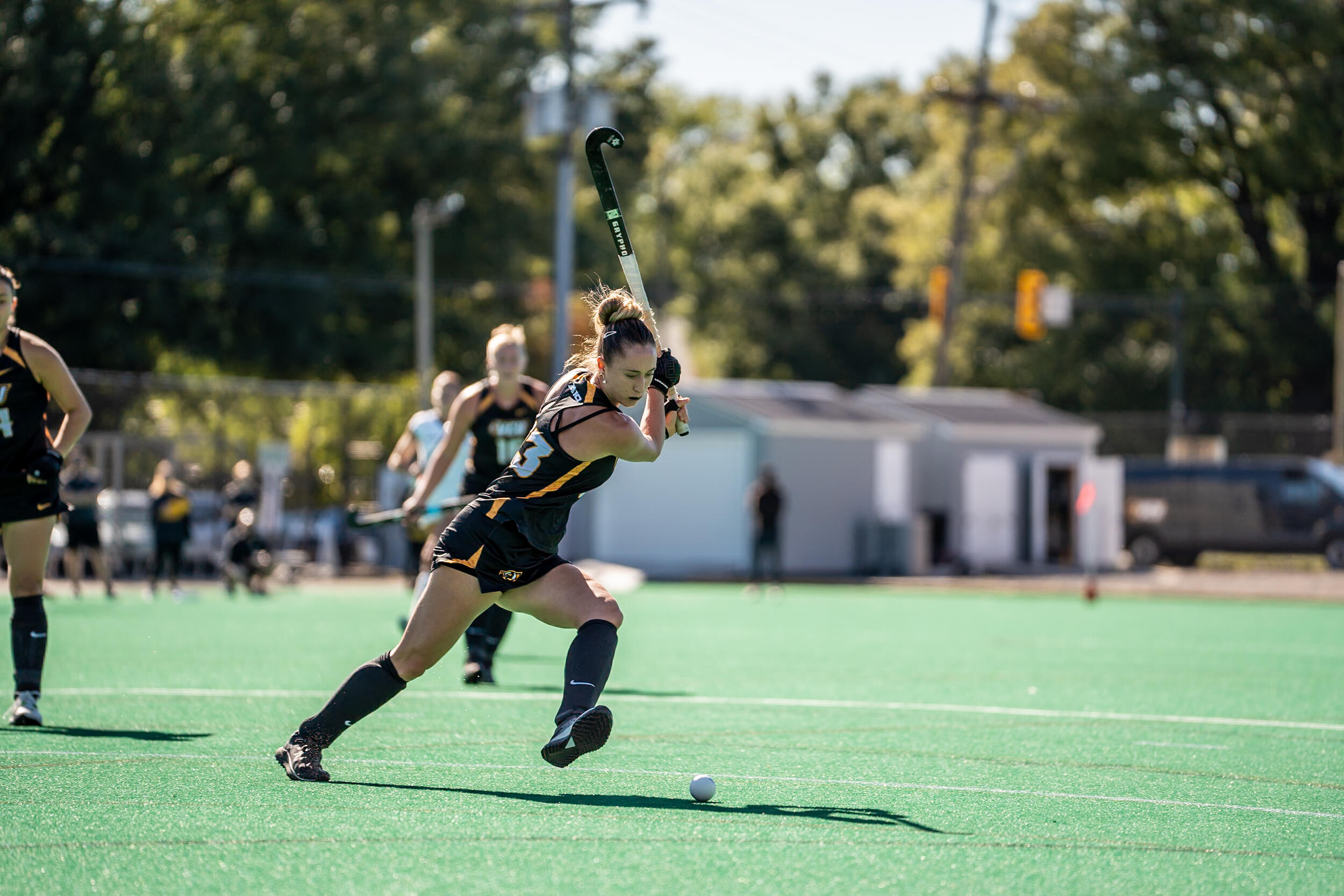 A photo of a woman running across a feild holding a field hockey stick above her head. She is looking down at a ball on the ground. 