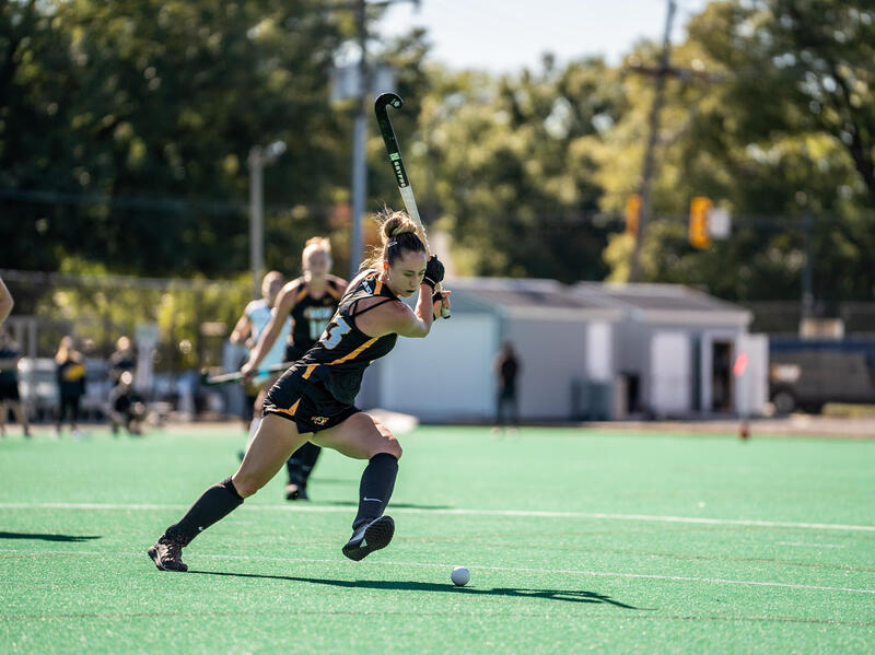 A photo of a woman running across a feild holding a field hockey stick above her head. She is looking down at a ball on the ground. 