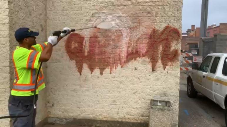 A man wearing a bright yellow shirt with orange and silver stripes using a power washer on the side of a building. 