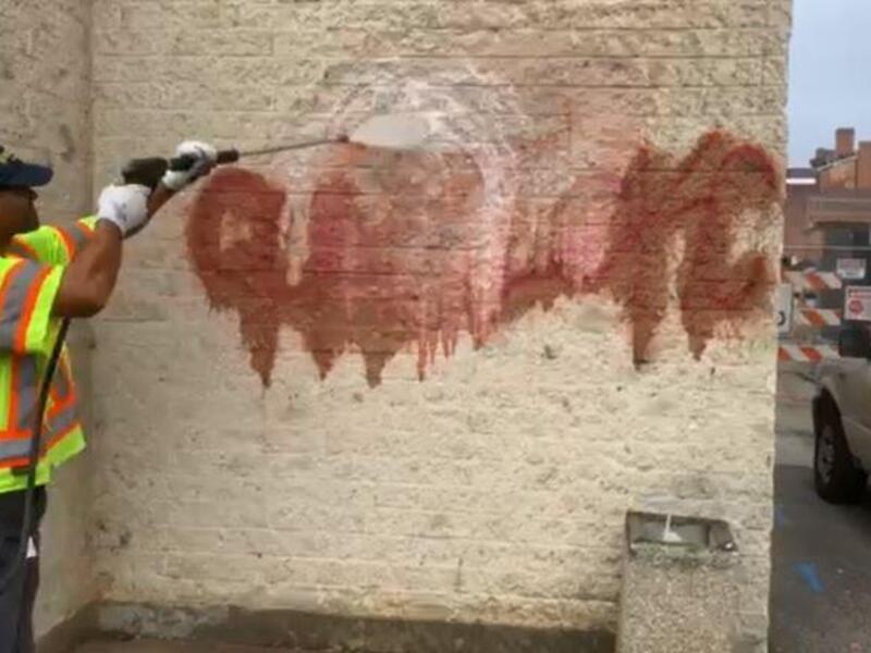 A man wearing a bright yellow shirt with orange and silver stripes using a power washer on the side of a building. 