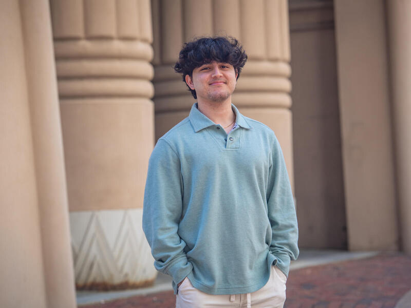 A man in a collared shirt smiling in front of columns with his hands in his pockets.