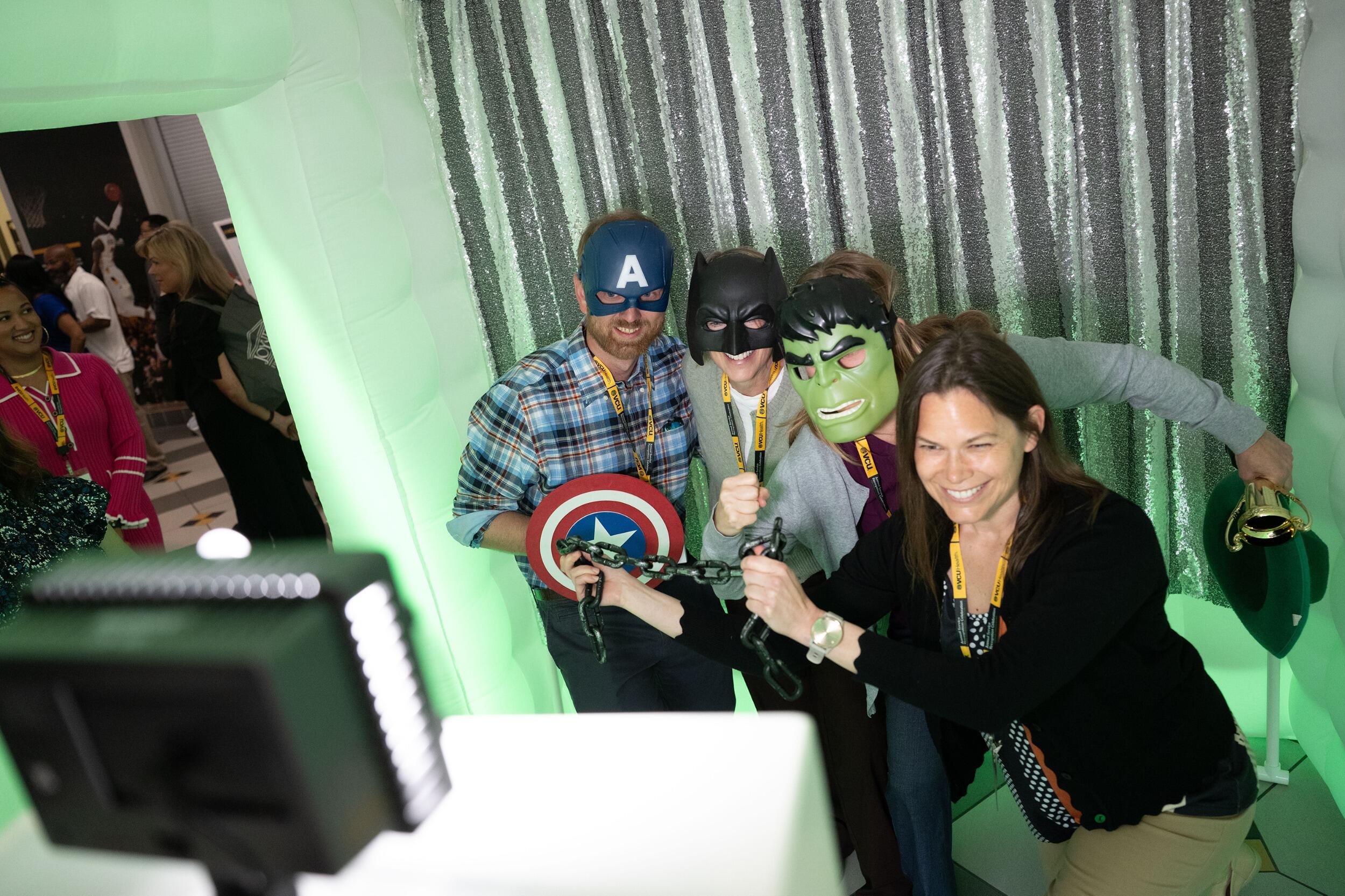 A photo of four people posing in a photo booth with superhero masks and props. 