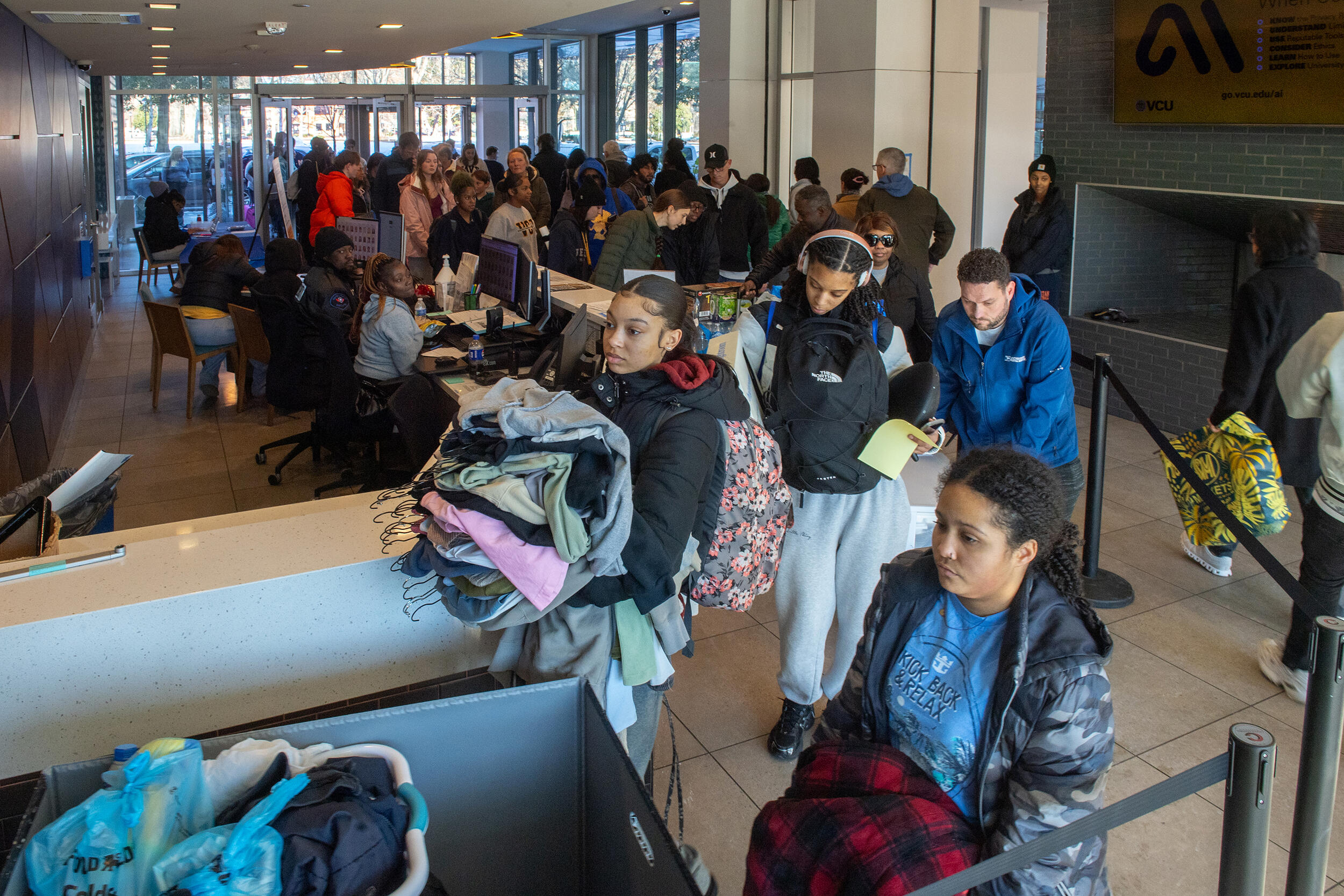 A photo of a dorm lobb filled with a corwd of people holding bags, clothing, and luggage. 