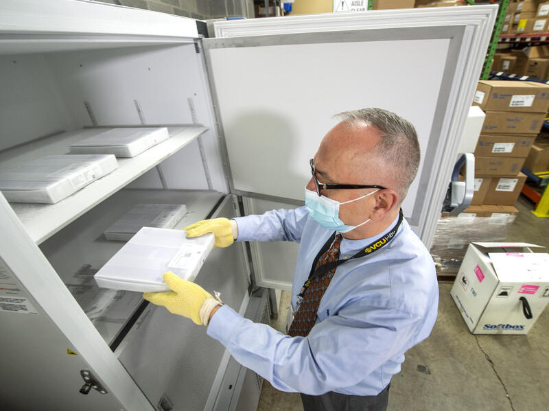 A person wearing a mask and gloves places a box containing vaccine vials into a freezer. 
