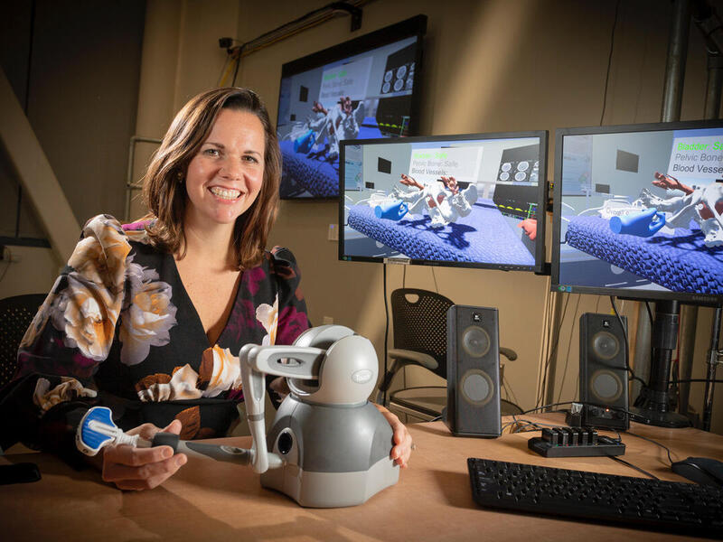 A woman holding a piece of robotics equipment sitting next to three computer screens with a 3D scenario on them